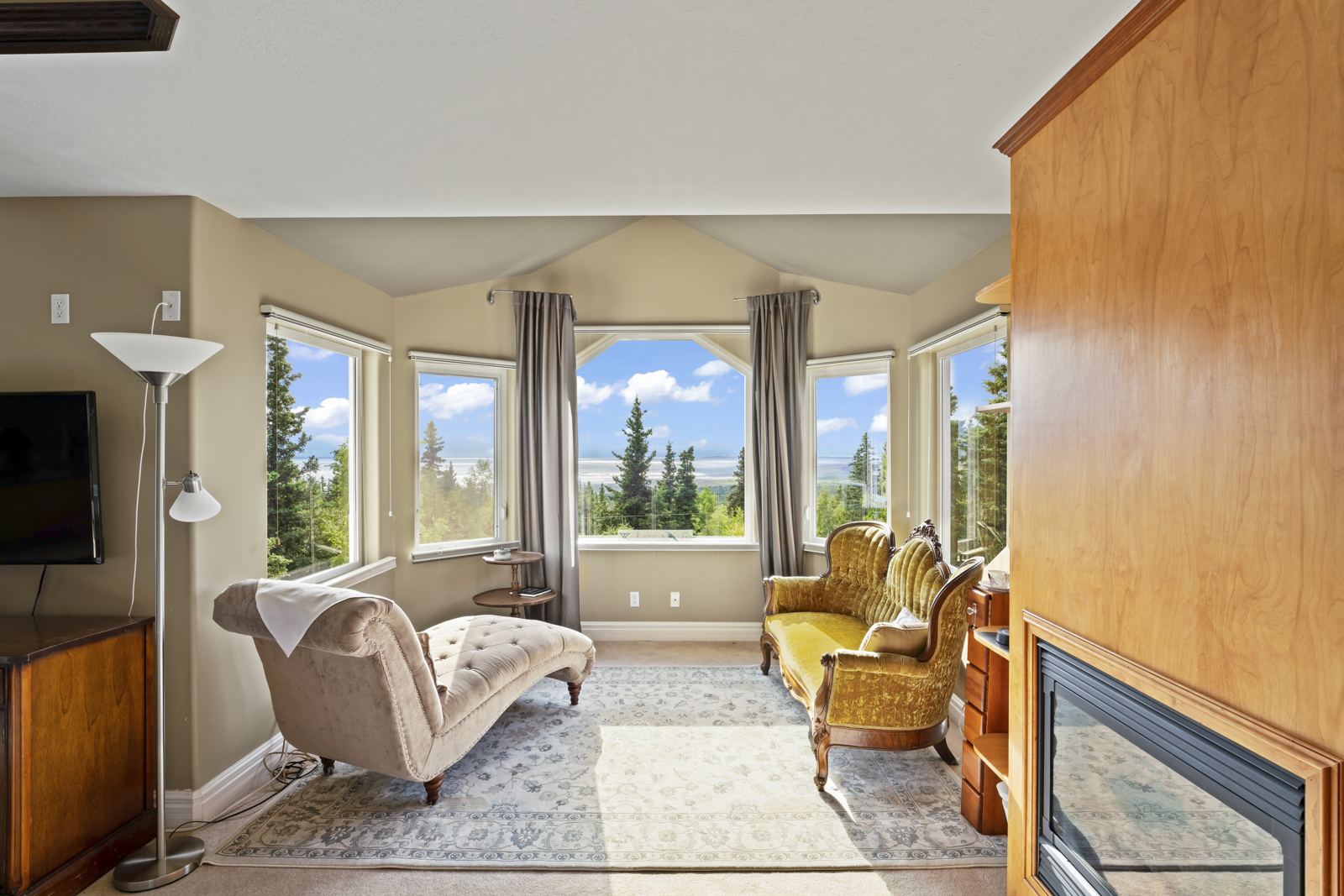 Primary suite sitting area at Hillside Haven: a chaise and fireplace beside bay windows looking over spruce to Cook Inlet